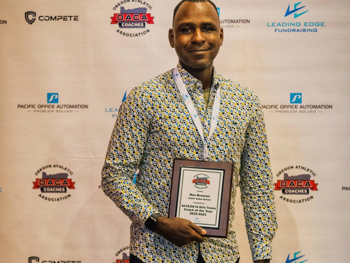 Ron Bronson holding a coach of the year plaque before an Oregon Athletic Coaches Association backdrop
